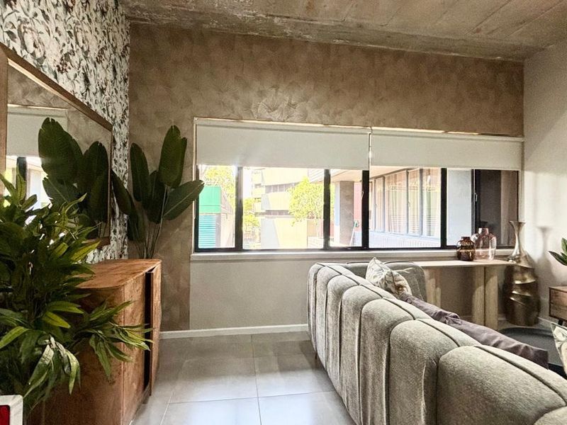 A well-maintained living room featuring an exposed concrete ceiling, large windows with roller blinds, and modern tiled flooring.