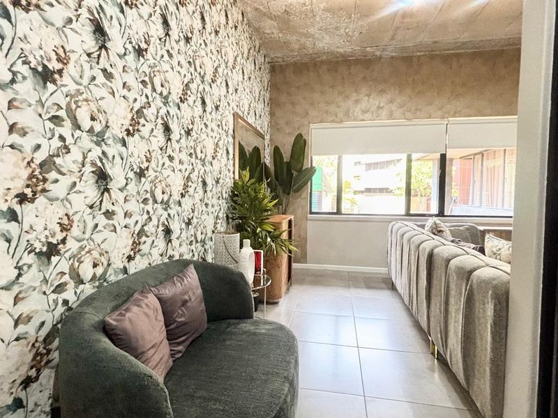 A living room featuring tiled flooring, large windows with roller blinds, and a distinctive floral wallpaper accent wall with an exposed concrete ceiling.