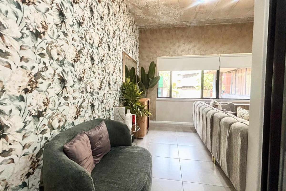 A living room featuring tiled flooring, large windows with roller blinds, and a distinctive floral wallpaper accent wall with an exposed concrete ceiling.