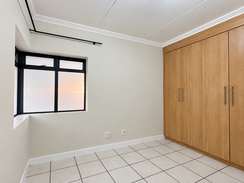 A bright, empty bedroom featuring neutral walls, tiled flooring, and extensive built-in wooden wardrobe storage with a black-framed window.