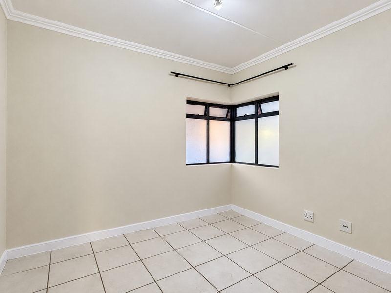 A bright, empty bedroom featuring neutral tiled flooring, a corner window with black frames, and classic ceiling cornicing.