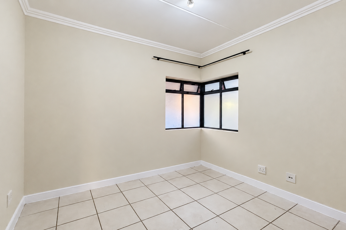A bright, empty bedroom featuring neutral tiled flooring, a corner window with black frames, and classic ceiling cornicing.