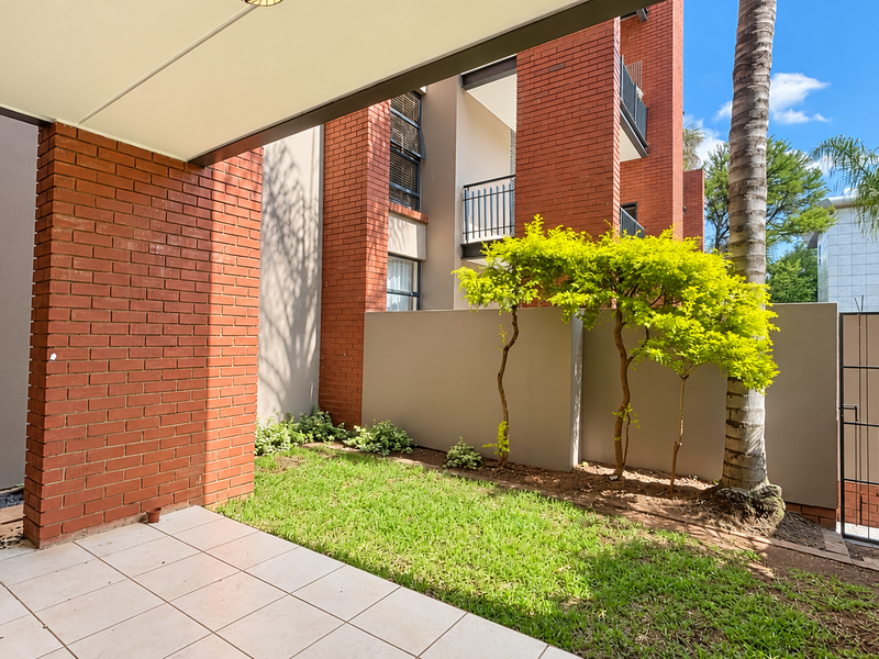 A private ground-floor patio featuring tiled flooring, brick architectural detailing, and direct access to a small landscaped garden area.