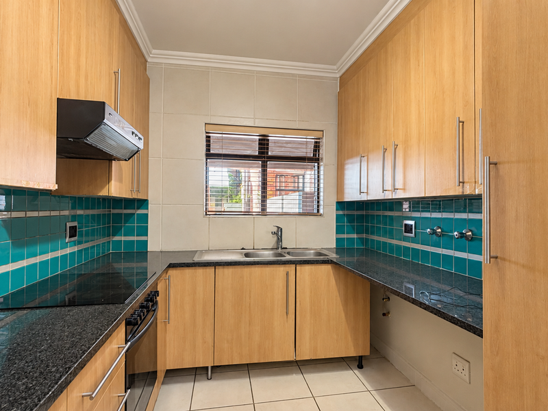 A clean, functional kitchen featuring light wood cabinetry, granite countertops, a built-in oven, electric hob, and a vibrant teal tiled backsplash.