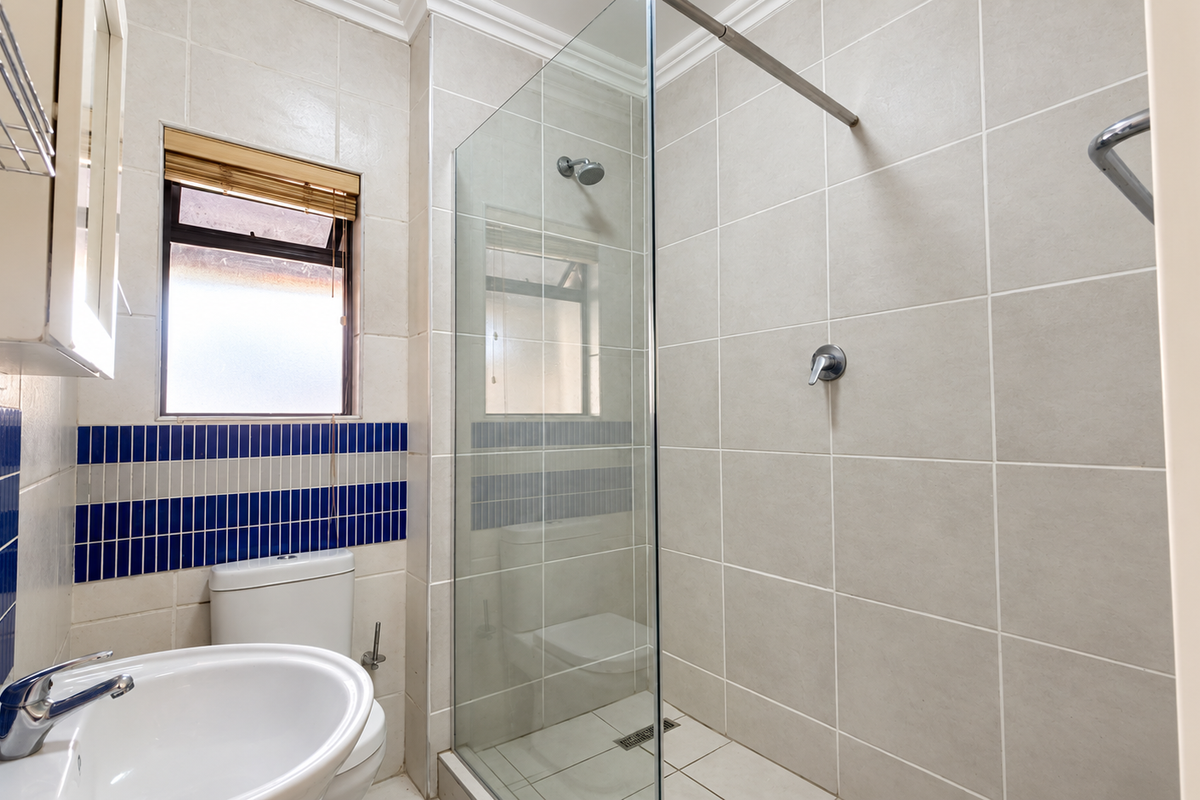 A functional bathroom featuring a glass-enclosed shower, ceramic basin, toilet, and neutral wall tiling with a decorative blue accent border.