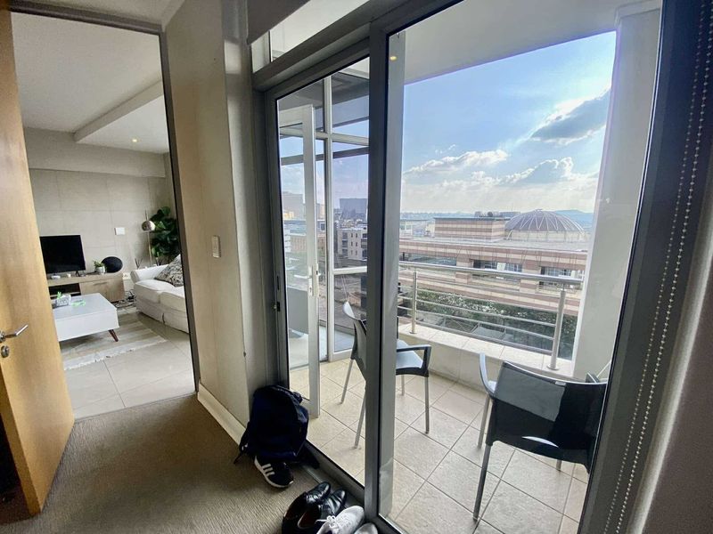 A balcony with tiled flooring and metal railings, offering an elevated urban view, accessible through a large sliding glass door from the living area.