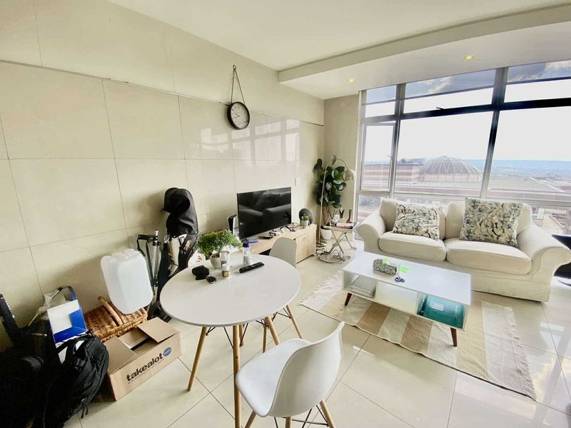 An open-plan living area featuring large floor-to-ceiling windows with natural light, tiled flooring, and a modern tiled feature wall.