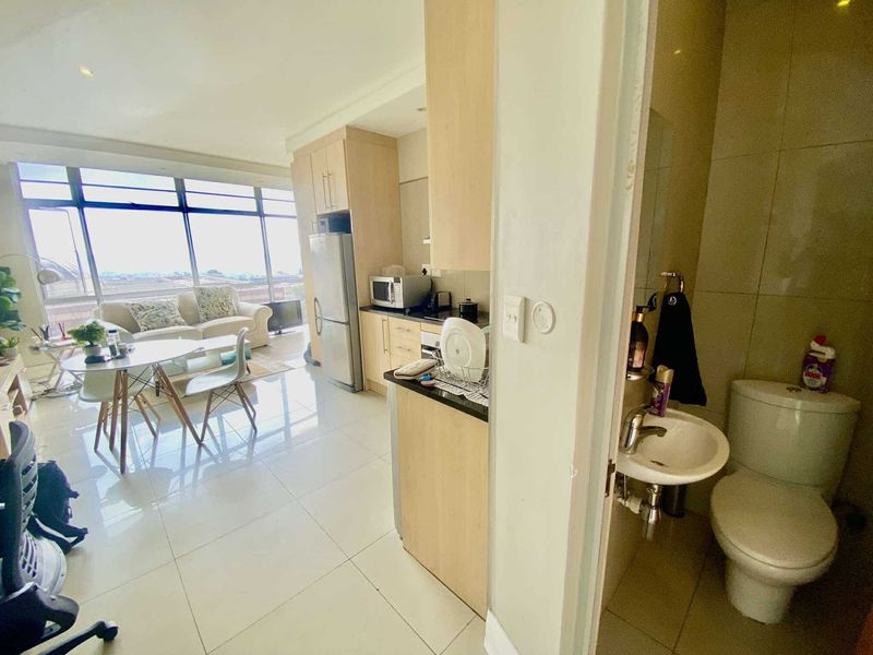 An open-plan apartment layout featuring a modern kitchen with granite countertops, tiled flooring, and large windows, adjacent to a compact bathroom.