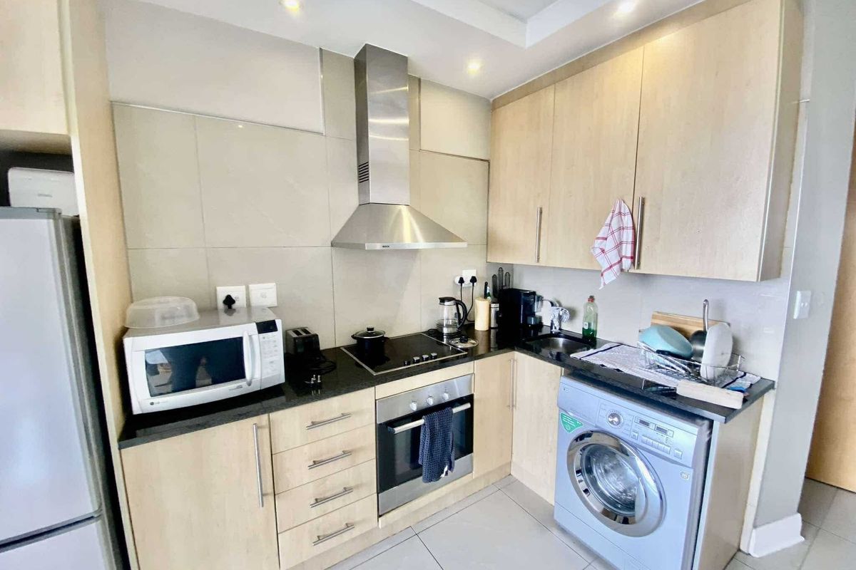 A compact, modern kitchen featuring light wood cabinetry, stone countertops, stainless steel appliances, and a built-in washing machine.