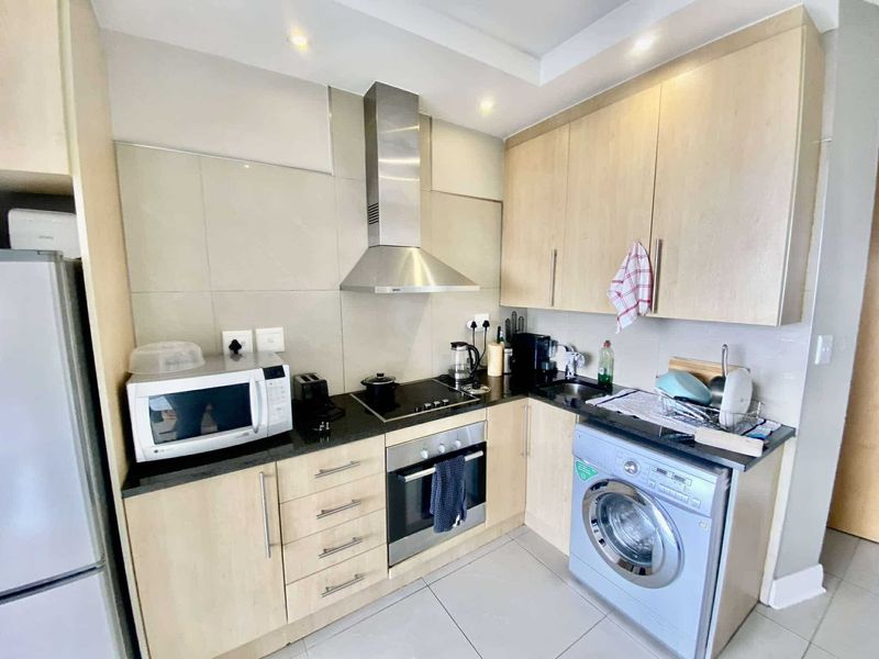 A compact, modern kitchen featuring light wood cabinetry, stone countertops, stainless steel appliances, and a built-in washing machine.