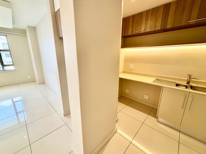 A clean, modern scullery area featuring built-in cabinetry, a stainless steel sink, and under-cabinet lighting, adjacent to an open-plan living space.