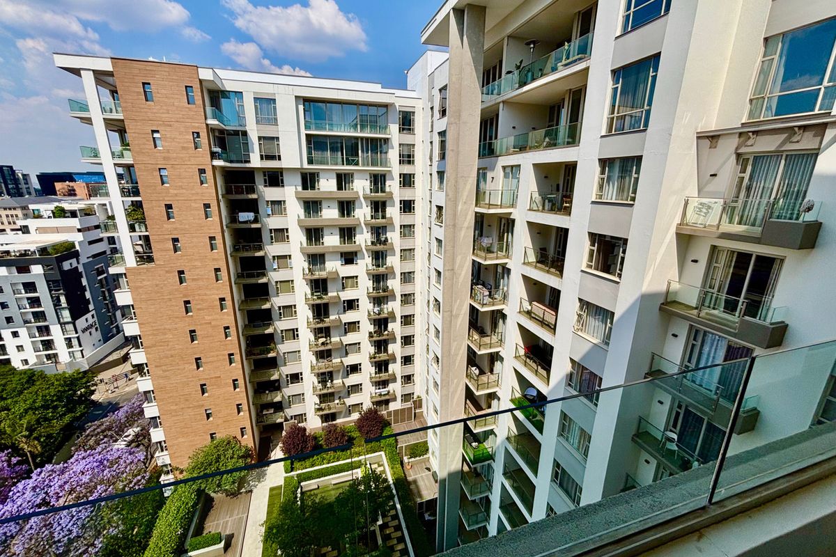 A high-angle view of a modern apartment complex featuring multiple balconies, glass balustrades, and a well-maintained landscaped courtyard area.