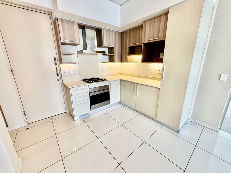 A modern, clean kitchen featuring light wood-tone cabinetry, stone countertops, integrated appliances including a gas hob and oven, and bright tiled flooring.