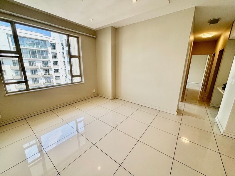 A bright and spacious empty living area featuring neutral tiled flooring, recessed lighting, and a large window providing natural light.