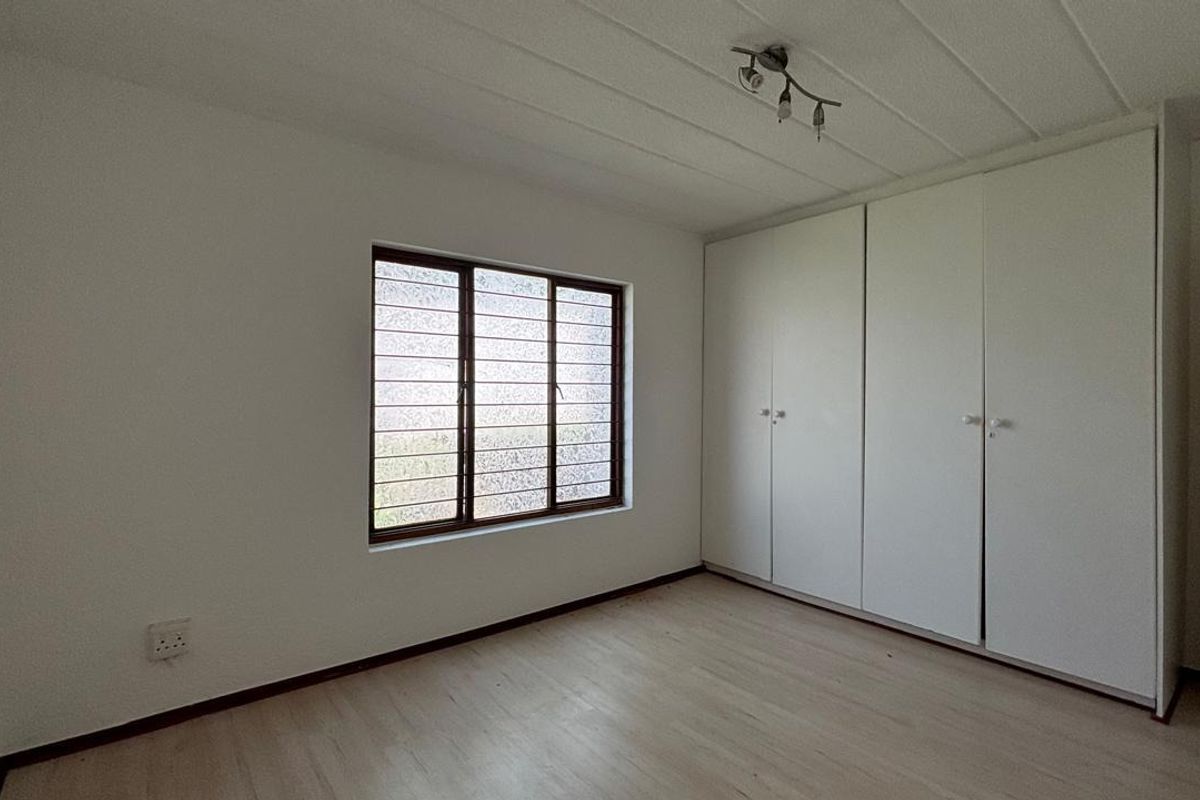 A bright, empty bedroom featuring built-in cupboards, laminate flooring, and a window fitted with security bars.