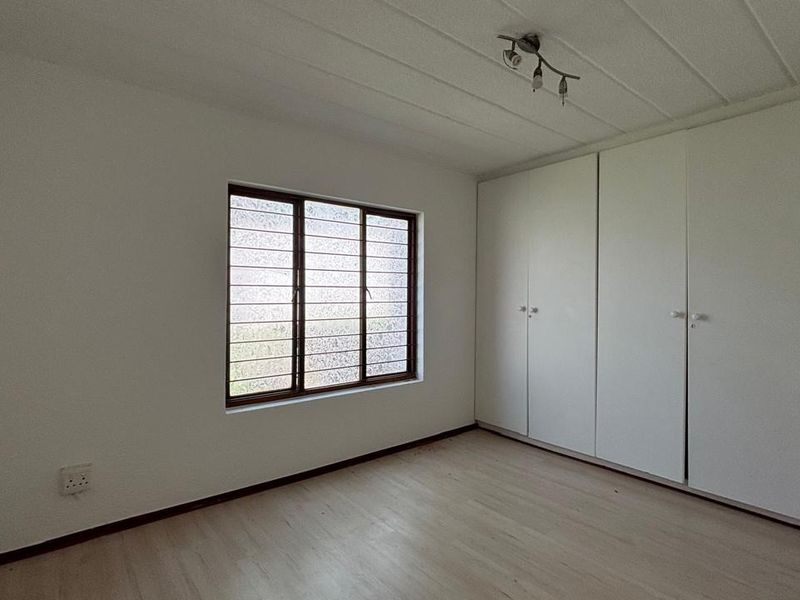 A bright, empty bedroom featuring built-in cupboards, laminate flooring, and a window fitted with security bars.