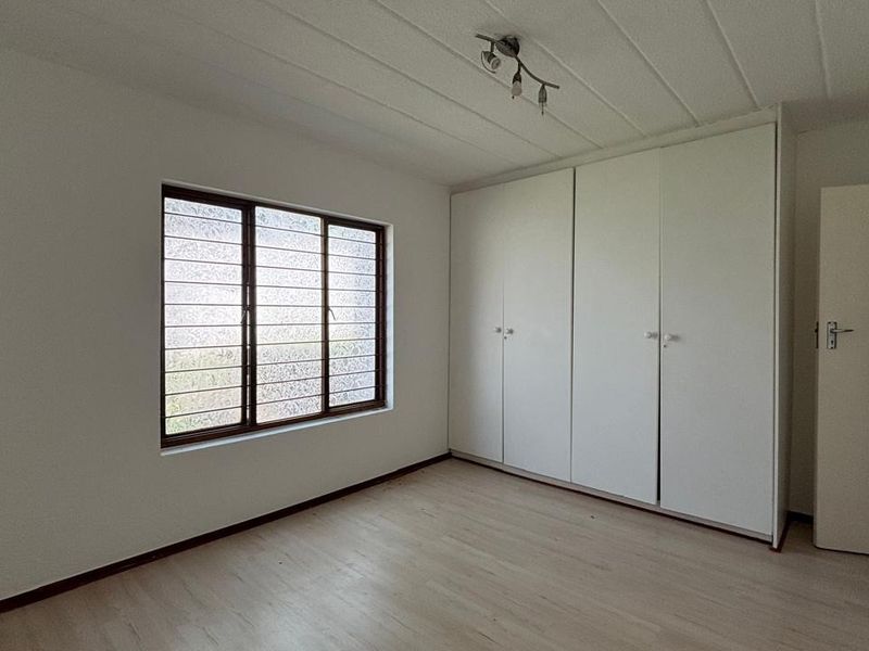 A bright, empty bedroom featuring built-in cupboards, laminate flooring, and a window fitted with security bars.