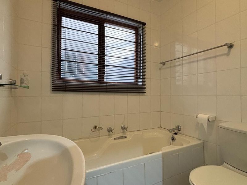 A functional bathroom featuring a built-in bathtub, pedestal basin, and toilet, finished with white wall tiles and a window for natural light.