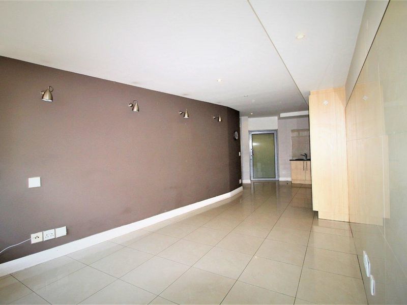 An open-plan living area featuring neutral tiled flooring, recessed lighting, and a modern wall-mounted spotlight arrangement, leading towards a kitchen and glass door entrance.