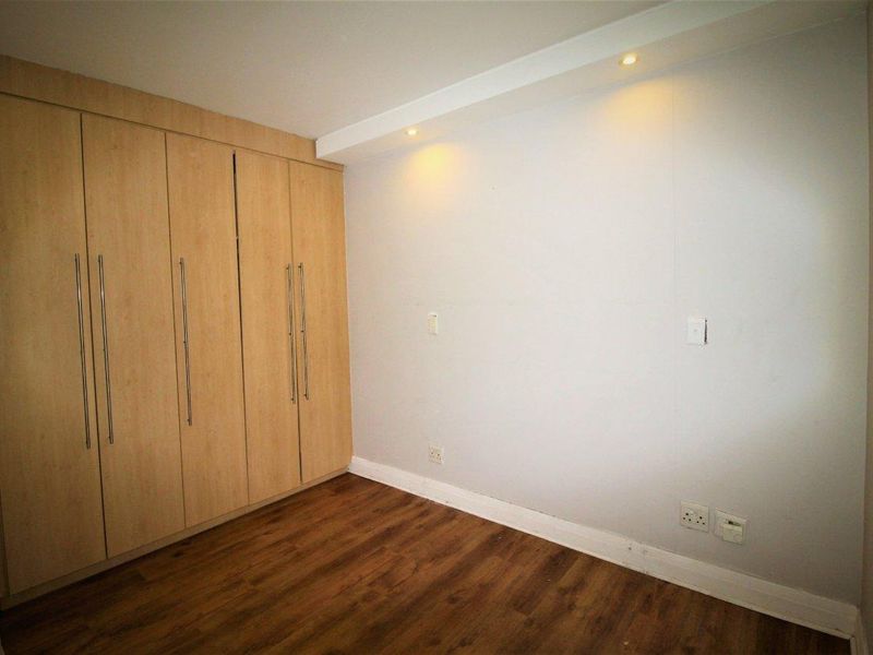 A clean, empty bedroom featuring light wood-finish built-in cupboards, warm laminate flooring, and recessed ceiling lighting.