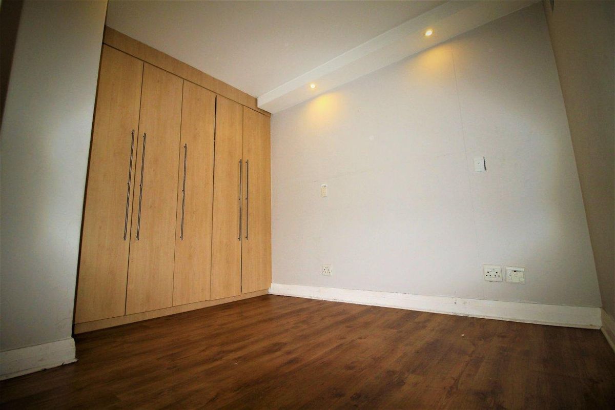 A clean, empty bedroom featuring light wood-finish built-in cupboards, warm laminate flooring, and recessed ceiling lighting.