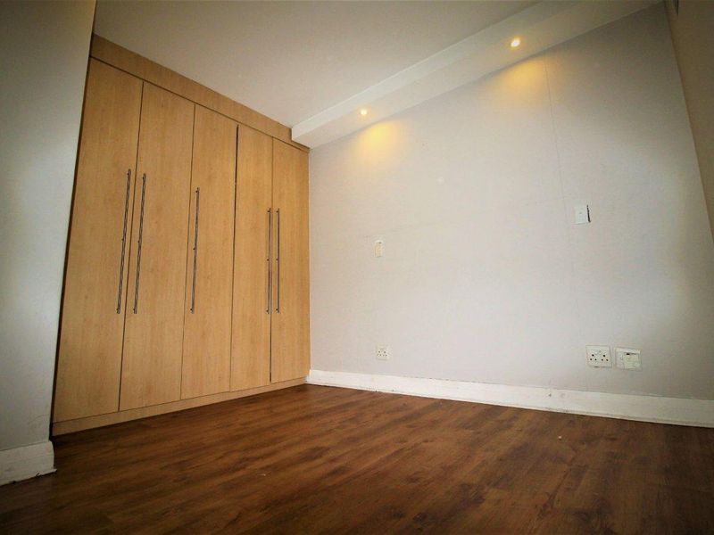 A clean, empty bedroom featuring light wood-finish built-in cupboards, warm laminate flooring, and recessed ceiling lighting.