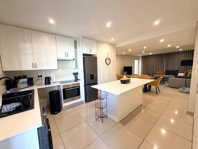 Modern open-plan kitchen and living area featuring white cabinetry, a central island with quartz countertops, integrated appliances, and sleek tiled flooring throughout.