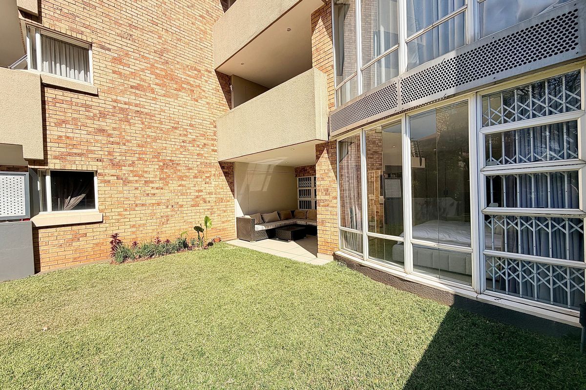 A rear view of the apartment building featuring a private lawn area, a covered patio space, and large windows with security bars.