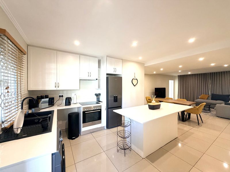 Modern open-plan kitchen and living area featuring sleek white cabinetry, a central island, integrated appliances, and neutral tiled flooring throughout.