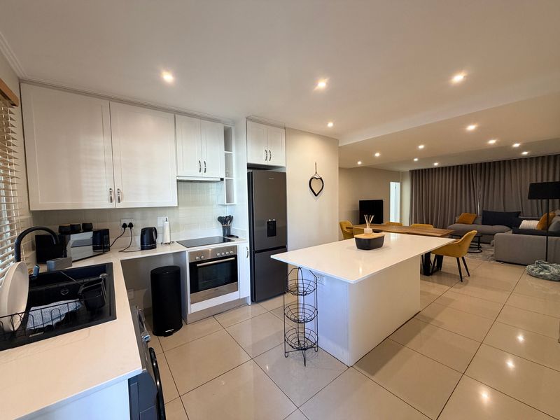 Modern open-plan kitchen and living area featuring a central island, sleek cabinetry, built-in appliances, and neutral tiled flooring throughout.