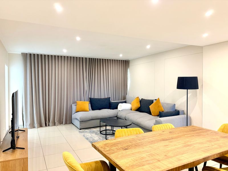 A bright and modern open-plan living and dining area featuring neutral tiled flooring, recessed downlighting, and floor-to-ceiling curtains.