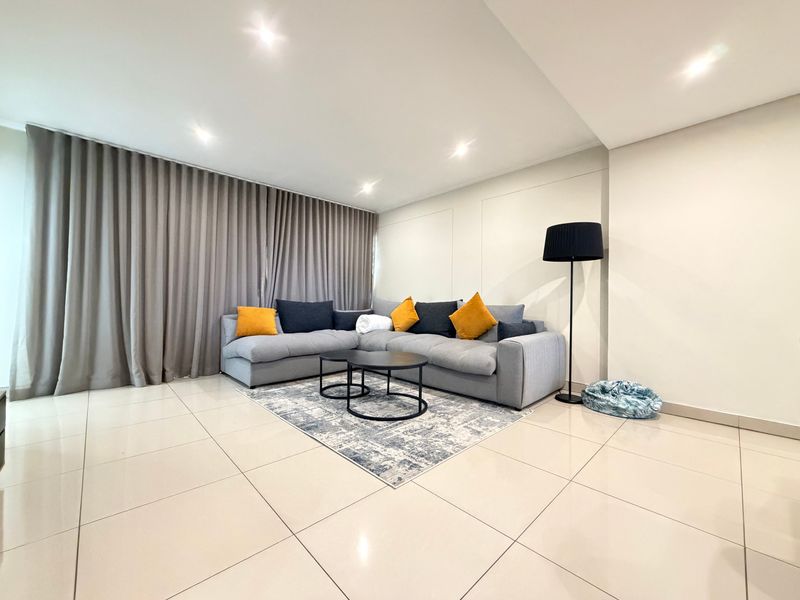 A bright and modern living room featuring neutral tiled flooring, recessed downlighting, and full-length window curtains.
