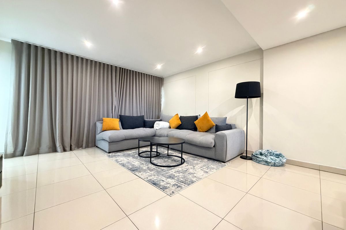 A bright and modern living room featuring neutral tiled flooring, recessed downlighting, and full-length window curtains.