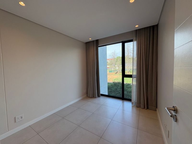 A bright, empty bedroom featuring modern tiled flooring, recessed downlighting, and a large floor-to-ceiling window with neutral curtains overlooking the exterior.