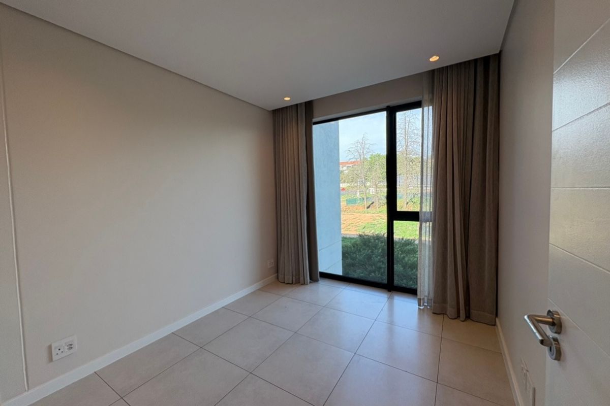 A bright, empty bedroom featuring modern tiled flooring, recessed downlighting, and a large floor-to-ceiling window with neutral curtains overlooking the exterior.