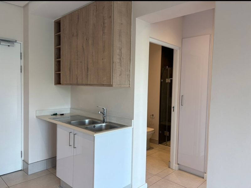 A compact kitchen area featuring modern wood-look upper cabinetry, white base cabinets, a stainless steel sink, and adjacent access to a bathroom.