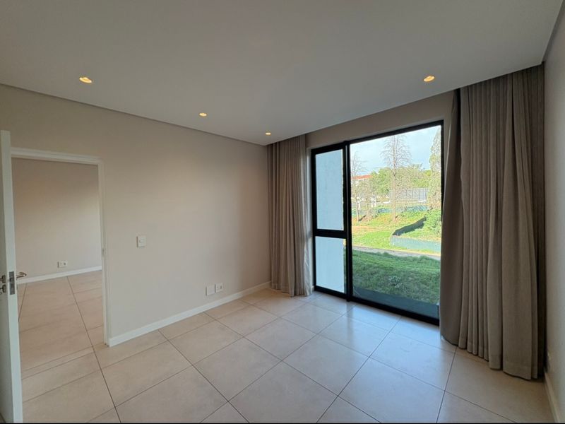 A bright, empty bedroom featuring neutral tiled flooring, recessed lighting, and a large sliding glass door with floor-to-ceiling curtains overlooking a green outdoor area.