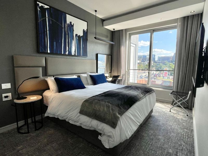 Modern bedroom featuring a large upholstered headboard, recessed ceiling lighting, and a sliding glass door leading to a balcony with city views.