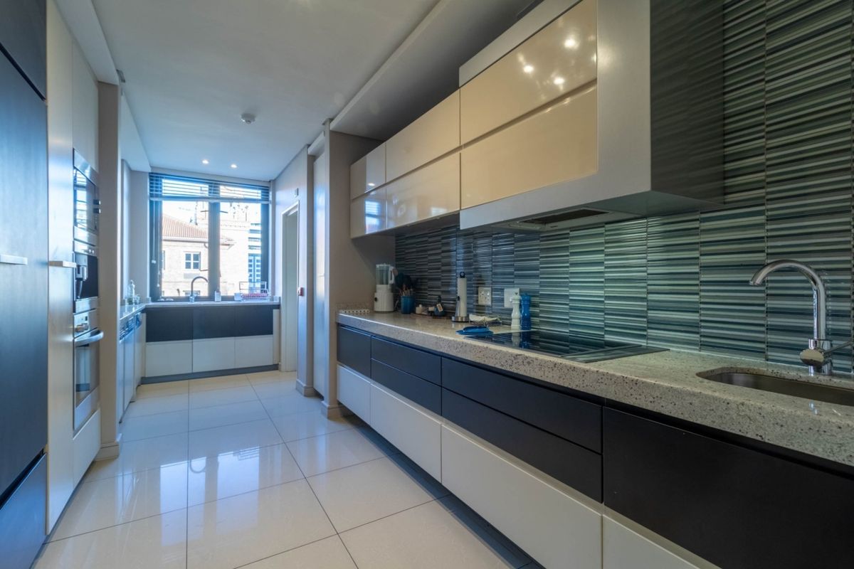 Modern galley-style kitchen featuring high-gloss cabinetry, stone countertops, integrated cooktop, and a tiled backsplash with a window overlooking the exterior.