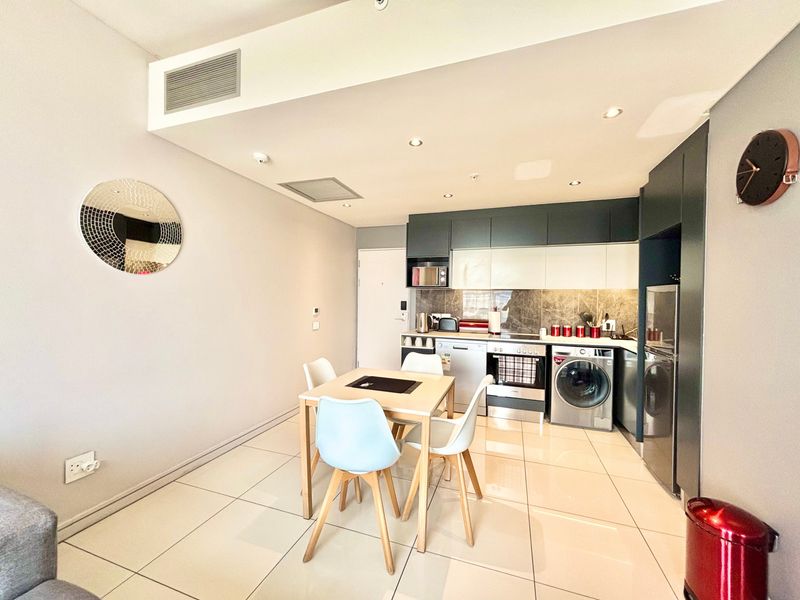 Modern open-plan kitchen and dining area featuring sleek built-in cabinetry, integrated appliances, tiled flooring, and recessed lighting in a contemporary apartment setting.