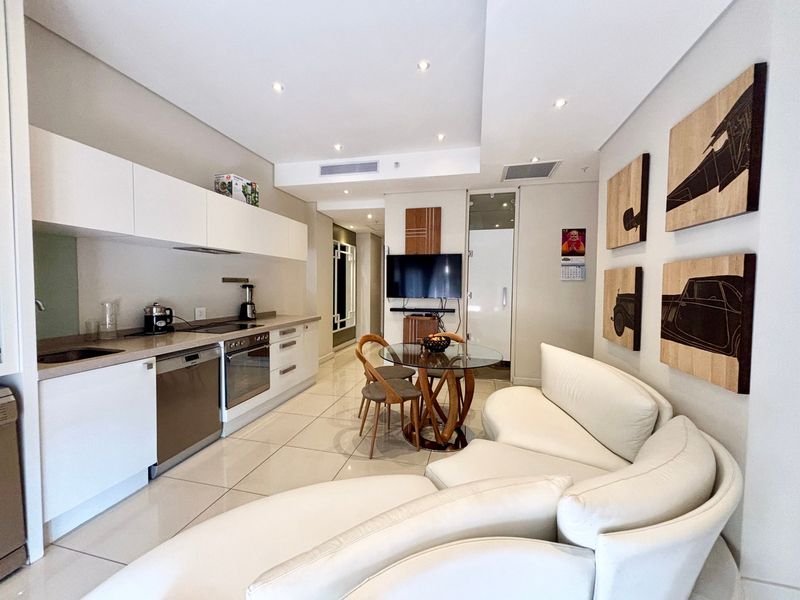 Modern open-plan apartment featuring a sleek kitchen with integrated appliances, tiled flooring, and recessed lighting, seamlessly flowing into the living and dining area.