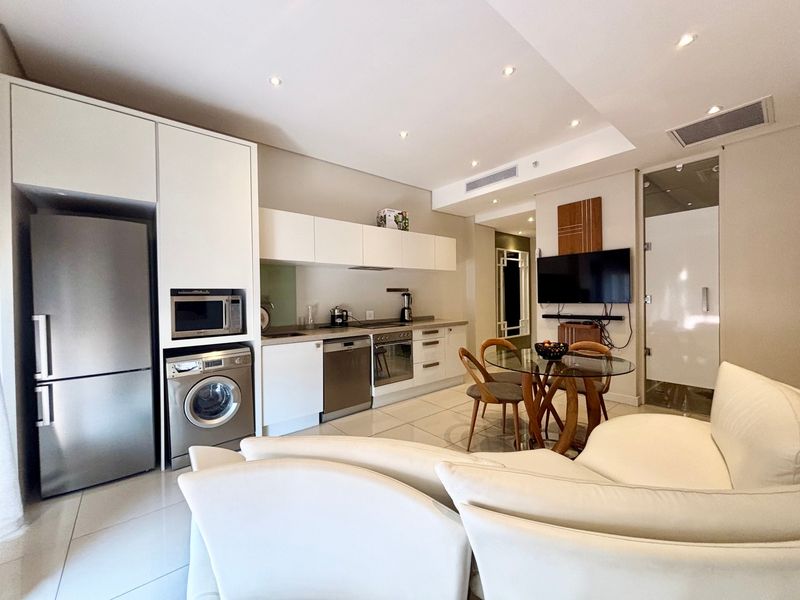 A modern open-plan living and kitchen area featuring sleek white cabinetry, integrated appliances, tiled flooring, and recessed lighting in a well-maintained apartment setting.