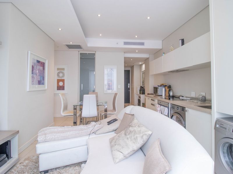 Modern open-plan living area featuring a sleek kitchen with integrated appliances, recessed lighting, and ducted air conditioning in a contemporary apartment setting.