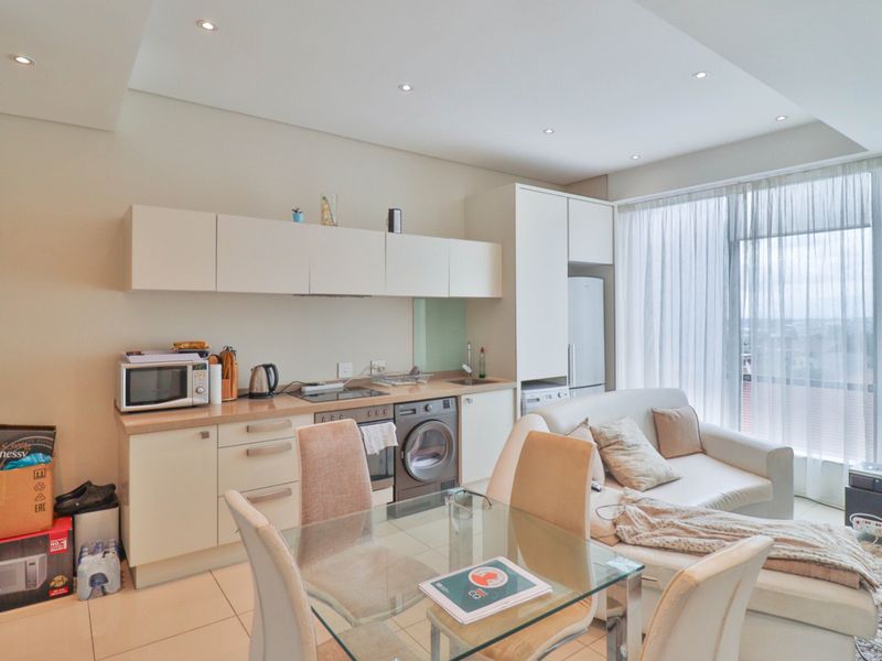 Modern open-plan apartment living area featuring a compact kitchen with built-in cabinetry, integrated appliances, and tiled flooring, leading to a bright living space.