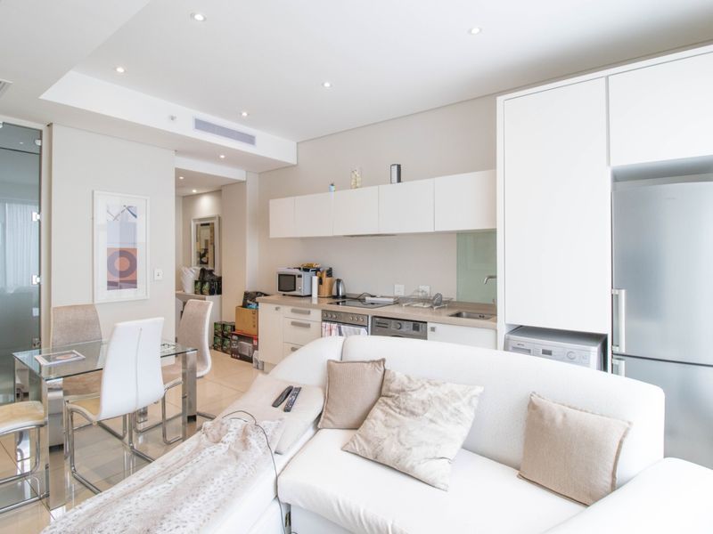 Modern open-plan living area featuring a compact kitchen with built-in cabinetry, integrated appliances, and recessed lighting, seamlessly flowing into the dining and lounge space.