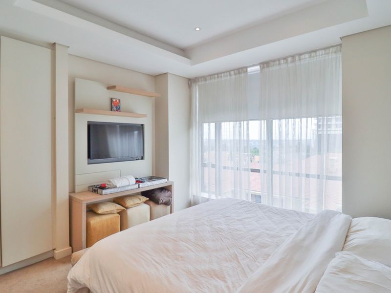 A well-maintained bedroom featuring a built-in wardrobe, custom media unit, bulkhead ceiling with recessed lighting, and large windows providing natural light.