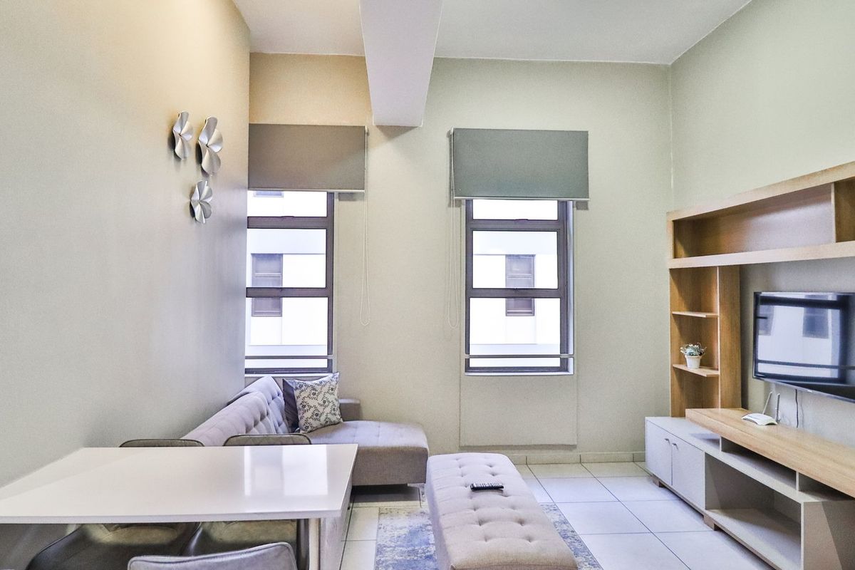 A well-maintained living room featuring tiled flooring, built-in wooden shelving, and large windows with roller blinds, offering a bright and functional living space.