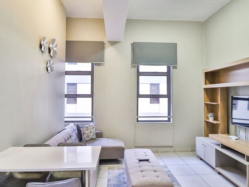 A well-maintained living room featuring tiled flooring, built-in wooden shelving, and large windows with roller blinds, offering a bright and functional living space.