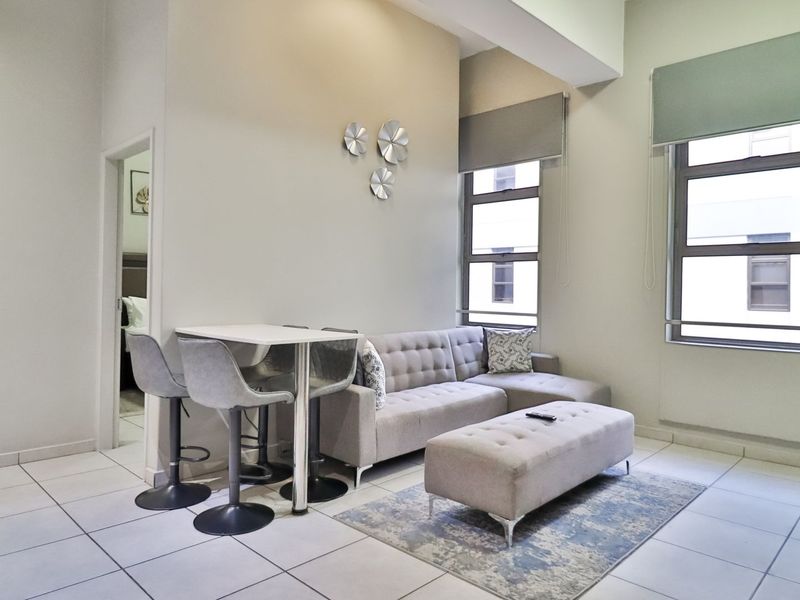 A bright and spacious living area featuring high ceilings, large windows with roller blinds, and neutral tiled flooring throughout.