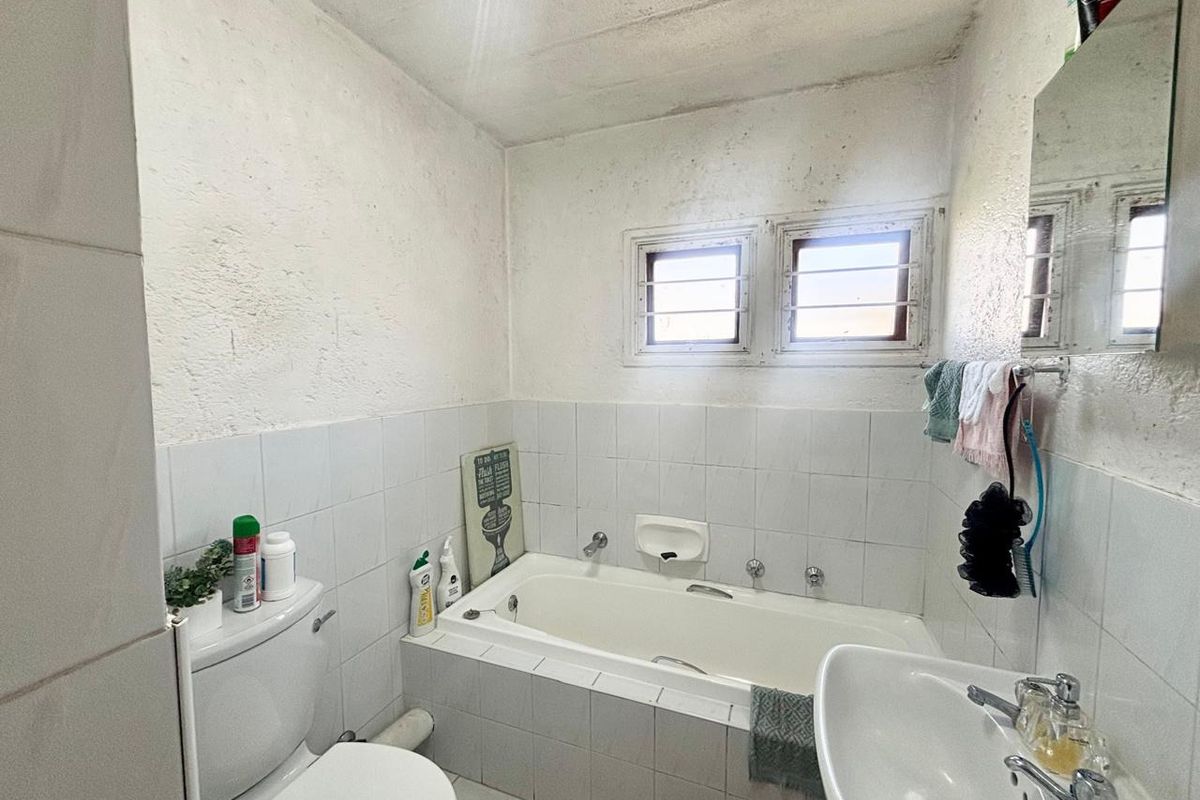 A functional bathroom featuring a built-in bathtub, pedestal basin, and toilet, with tiled finishes and security bars on the windows.